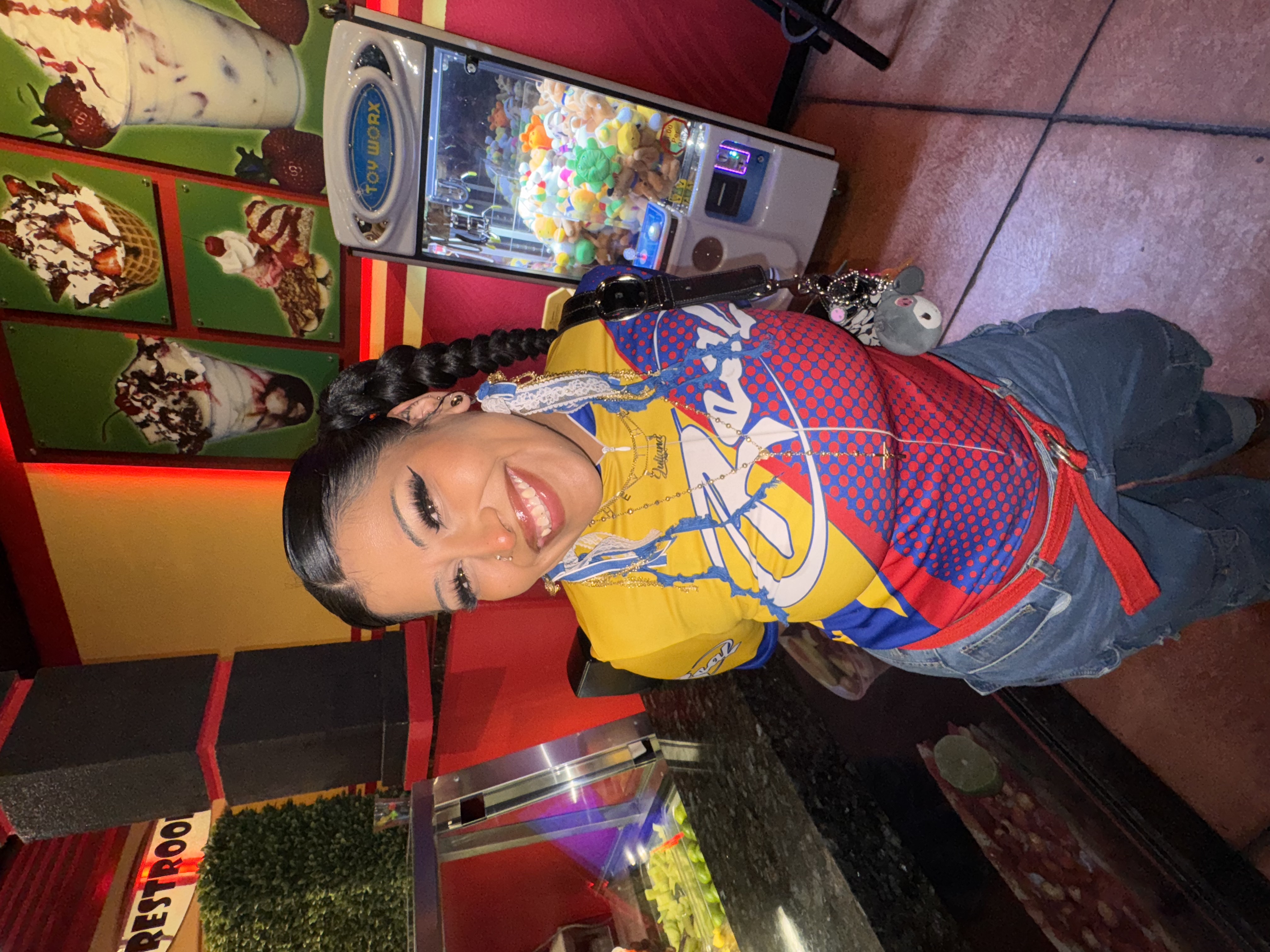 yessica smiles at the camera while standing with their arms behind their back. they have braids, a bright colorful shirt on, and arcade games behind them.