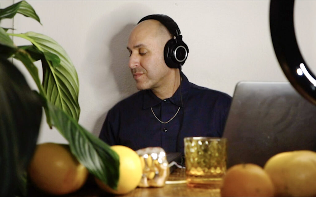 Joe with his eyes closed, head turned to his left, with headphones on. in front of him is a table with a orange glass, a laptop, oranges, and green leaves from a house plant