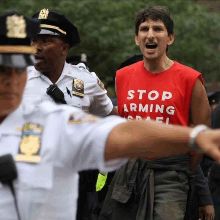 Morgan is chanting while wearing a red shirt that says "stop arming israel" and is surrounded by cops who are arresting them and pointing to the right.