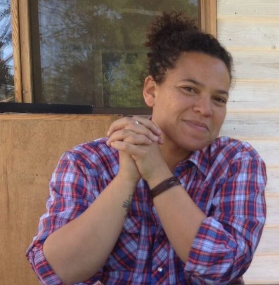 Rachel wearing a plaid purple shirt, looking at the camera with her hands clasped and elbows leaning on a table covered with scrabble board and parts.