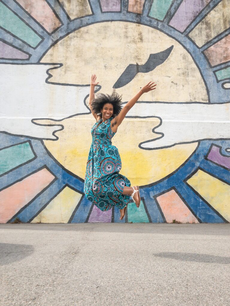 Alexis jumping while wearing a turquoise dress and her hands in the air, in front of a colorful mural of rainbow colored sunrays and stylized clouds
